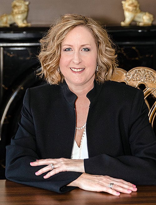 Professional headshot of Jennifer Martinez - woman with shoulder-length blonde wavy hair wearing a black blazer over white top with pearl necklace, positioned in three-quarter pose with hands on wooden surface, displaying rings on fingers, in elegant interior setting with decorative golden lion figurines and ornate carved chair in background