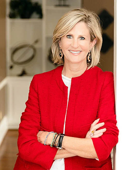 Professional portrait of Sarah Mitchell - woman with shoulder-length blonde hair wearing striking red blazer with white trim details over white top, arms crossed in confident pose, elegant jewelry including dark drop earrings and multiple bracelets, photographed in upscale interior with cream walls and decorative furnishings in background